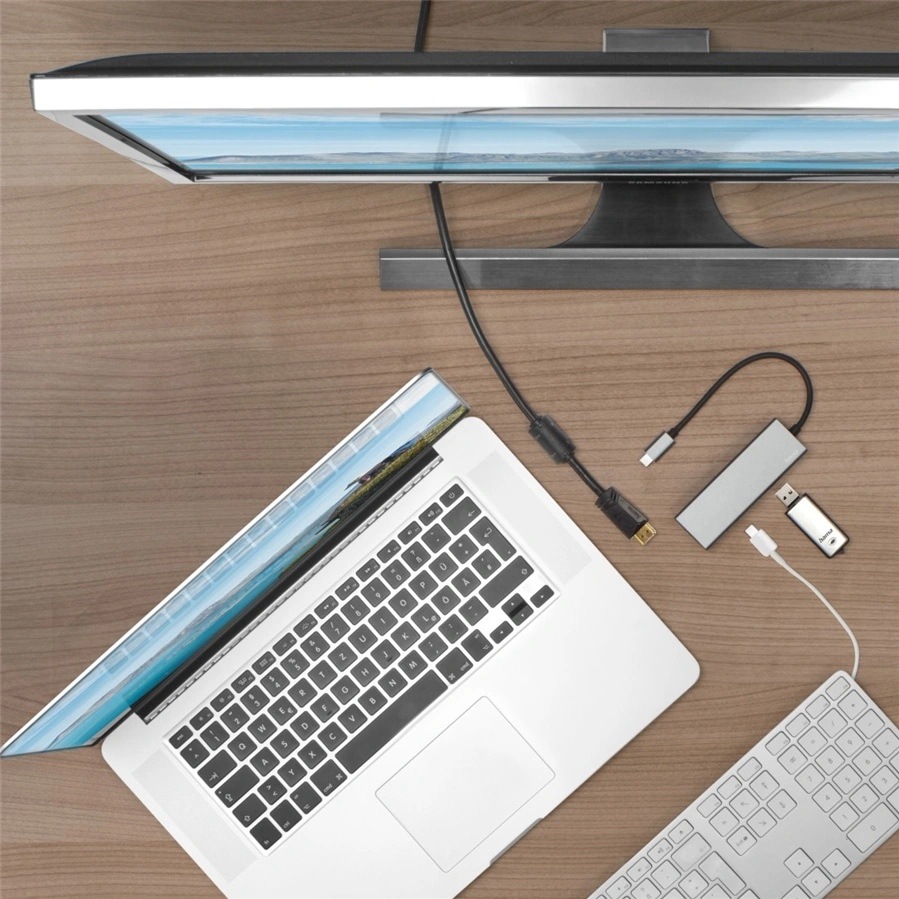 USB Hub Hama Multiport sivý Na obrázku je pracovný stôl z pohľadu zhora. Uprostred leží otvorený strieborný notebook, hore je veľký monitor a vpravo dole biela klávesnica. Vedľa notebooku je položený šedý USB-C hub Hama s krátkym káblom a niekoľko ďalších káblov a malých adaptérov. Fotka ukazuje bežné použitie hubu v pracovnom prostredí pri pripojení notebooku k monitoru a ďalšiemu príslušenstvu.