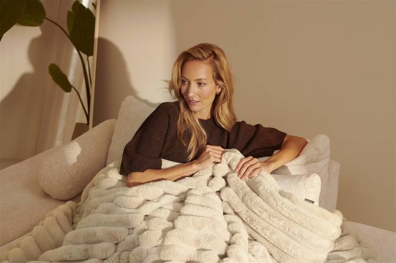 Vyhrievacia deka Duux DXOB01 Yentl Cuddle 200 x 200 cm béžová Žena pololeží v širokom kresle v teplom večernom osvetlení, zababušená do mäkkej béžovej vyhrievacej deky Duux Yentl, v pozadí vysoká izbová rastlina a pokojná domáca atmosféra