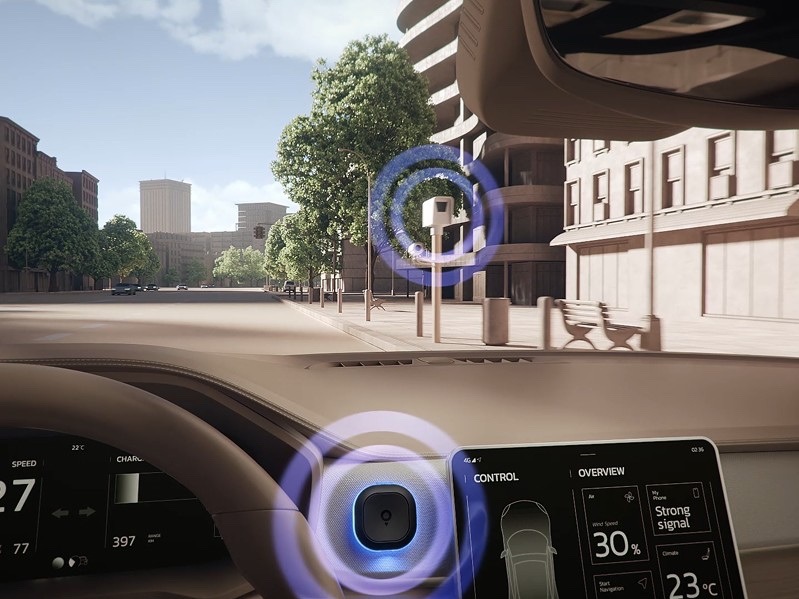 Pohľad z interiéru automobilu na mestskú ulicu. Cestovný asistent TomTom Tom je upevnený na palubnej doske vedľa dotykového displeja a modro svietiacim LED krúžkom upozorňuje na rýchlostnú kameru pred vozidlom.