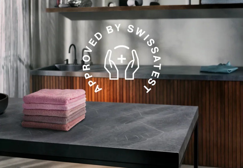 Stôl s kôpkou ružovej bielizne. V pozadí linka s drezom. V popredí symbol pre certifikáciu Swissatest.