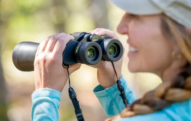 Usmievavá pani drží v ruke ďalekohľad Nikon Action 7×50 a pozerá sa pred seba na krajinu.