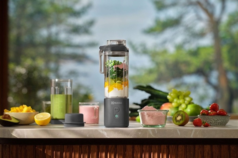 Stolný AKU mixér Electrolux E4PB1-2CG na kuchynskej linke. Vnútri mixéra je zmes pre smoothie. Na linke v pozadí sú misky s ovocím.