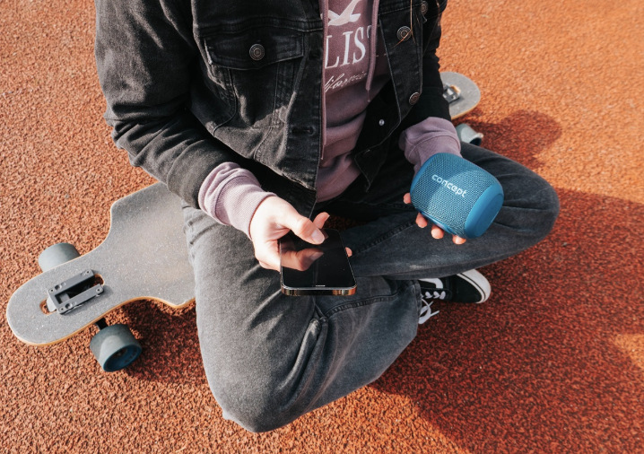 Prenosný reproduktor Concept SoundJoy BR5012 modrý Užívateľ drží modrý reproduktor Concept SoundJoy BR5012 a telefón pri posedení pri longboarde