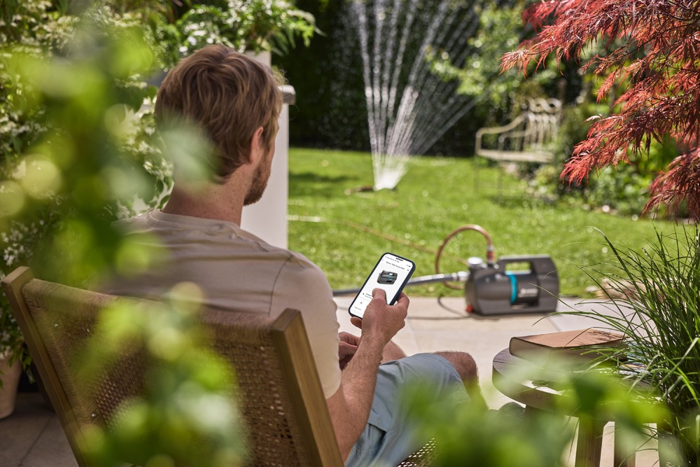 Vodáreň Gardena 5000 Silent Comfort 09072-20 Muž ovláda zavlažovanie záhrady pomocou mobilného telefónu, čerpadlo je pripojené k postrekovaču.