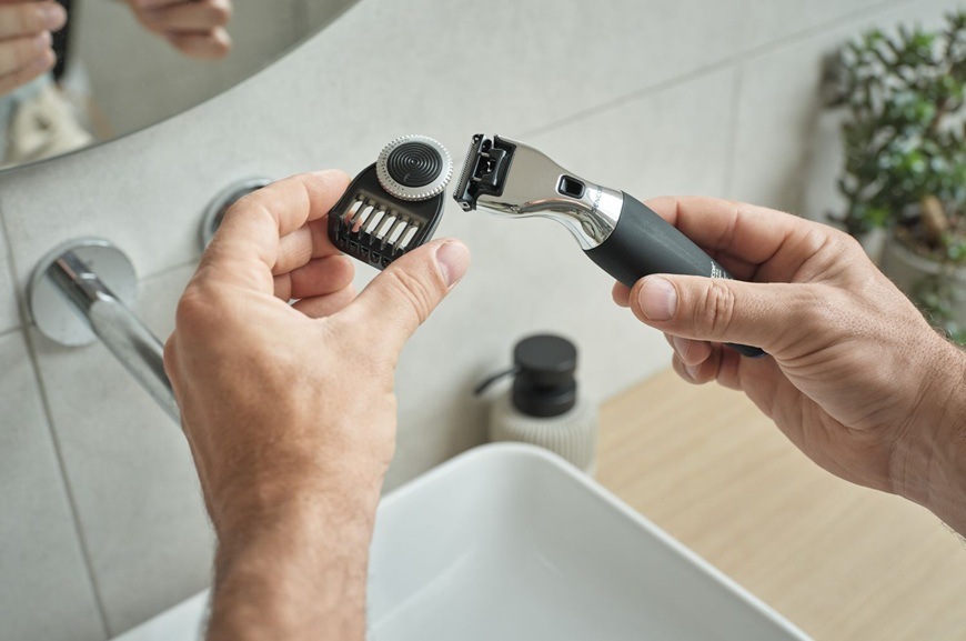 Muž drží elektrický zastrihávač Sencor SHP 0460BK a nasadzuje naň vymeniteľný nadstavec na úpravu fúzov. Scéna sa odohráva v kúpeľni nad umývadlom, kde sú viditeľné kúpeľňové doplnky a zrkadlo.
