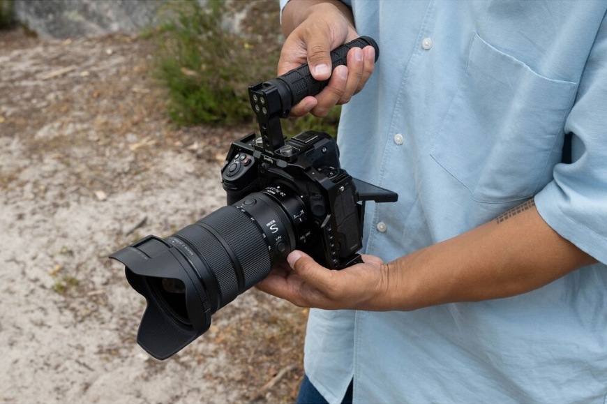 Detail objektívu NIKKOR Z 24 – 70 mm f/2.8 S II upevneného k fotoaparátu v rukách fotografa