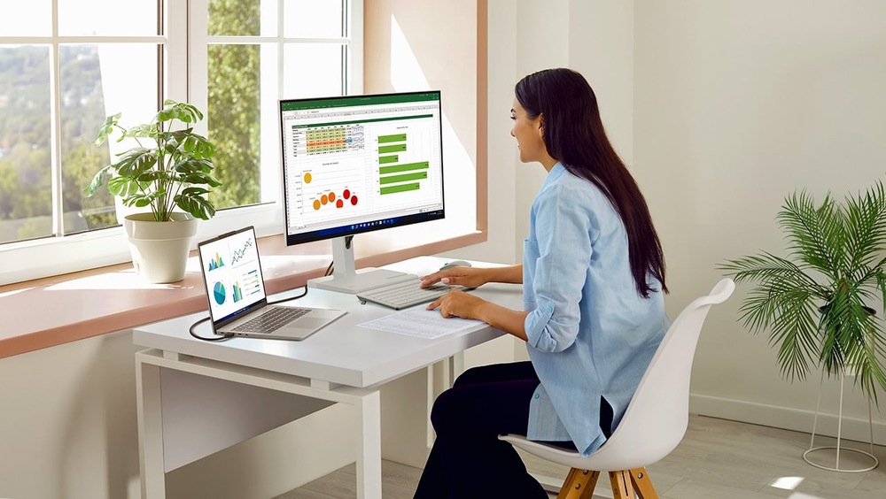 Monitor Lenovo L27h-4A sivý Žena pracuje pri stole s notebookom a monitorom, na ktorom sú zobrazené tabuľky a grafy. Svetlá miestnosť s veľkými oknami, rastlinami a pokojnou, sústredenou atmosférou.