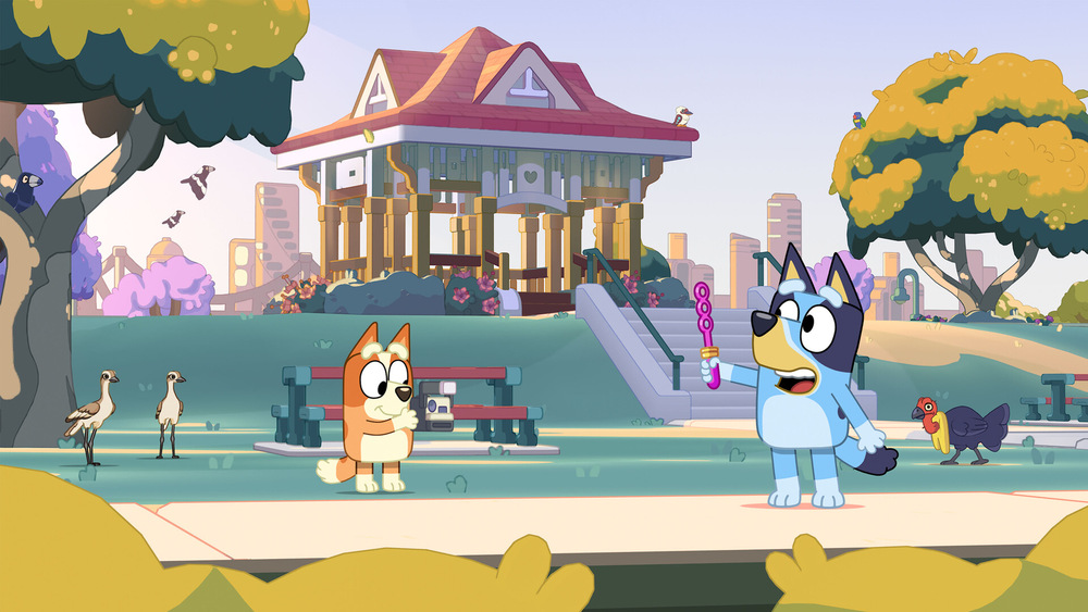 Bluey a Bingo stoja v slnečnom parku s altánkom a stromami. Bluey drží bublifuk. V pozadí sú vtáky a sliepky, čo celej scéne dodáva hravú atmosféru.