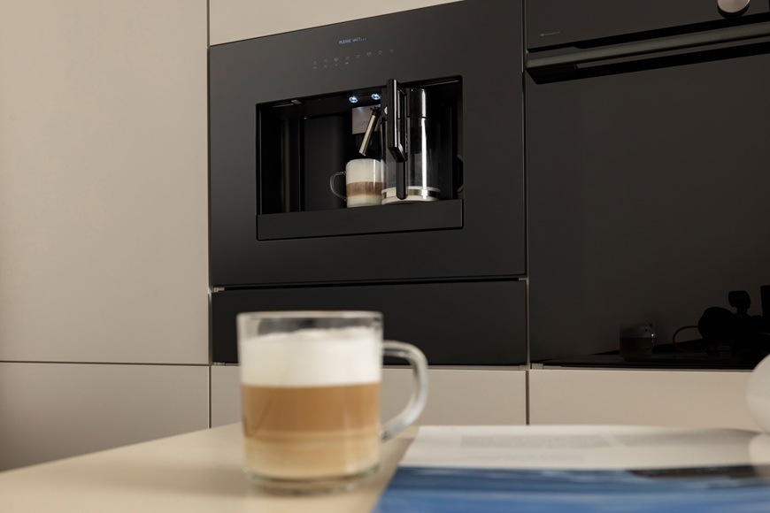 Espresso Gorenje G800 CM4G8BG čierna Na stole je položený hrnček s cappuccínom. V pozadí je zabudovaný kávovar Gorenje CM4G8BG, ktorý práve dovaril kafé s bohatou penou.