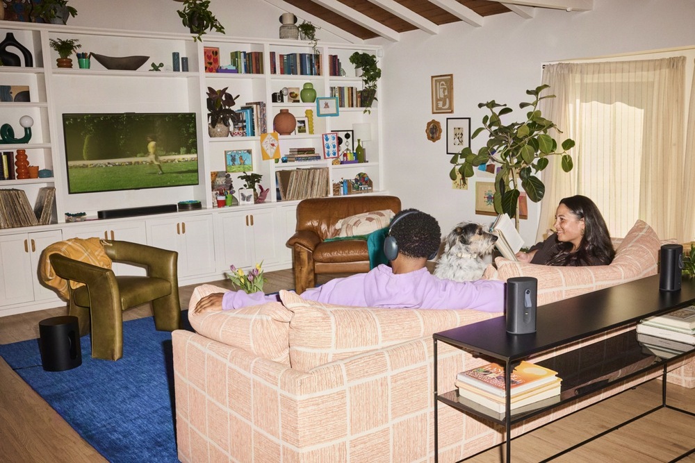 Soundbar SONOS Beam Ultra čierny Pár relaxuje na ružovej vzorovanej pohovke vo svetlom obývacej izbe s dreveným stropom, vysokou bielou knižnicou, zelenými izbovými rastlinami a čiernym reproduktorom Sonos na konzolovom stolíku.