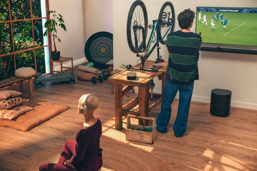 Muž opravuje bicykel a sleduje futbal v televízii, pod ktorou sa nachádza subwoofer JBL BAR 1000 MK2
