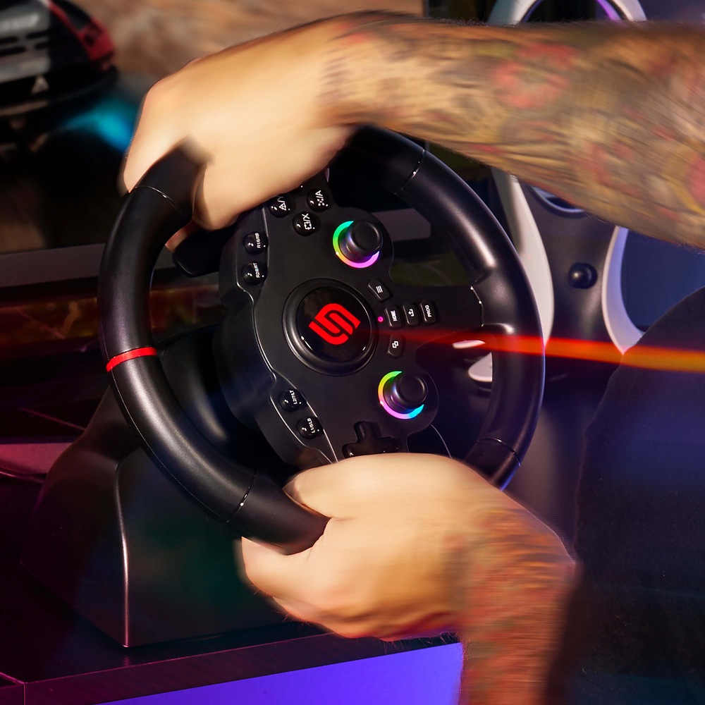 Volant SUPERDRIVE 220 RS-5 čierny Tetované ruky zvierajú herný volant s LED podsvietením analógových páčok. V pozadí je rozmazaná obrazovka závodnej hry, ktorá evokuje vzrušenie a rýchlosť.