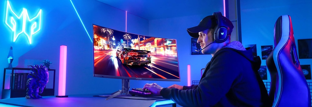 Hráč s nasadenými slúchadlami a šiltovkou sedí pri modernom hernom zariadení v modro osvetlenej miestnosti. Na veľkom zakrivenom monitore vidíme scénu závodnej hry.
