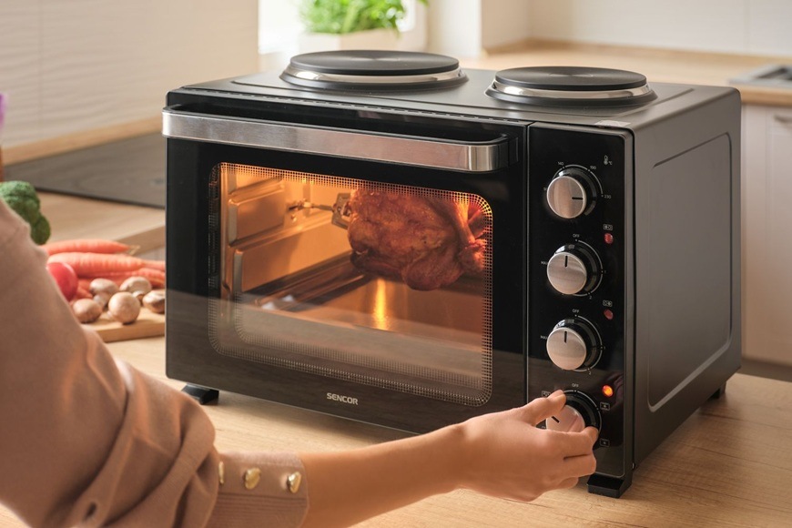 Mini rúra Sencor SEO 3500BK s otočným grilom pečie celé kurča, zatiaľ čo používateľ nastavuje režim pečenia pomocou otočných ovládačov v modernej kuchyni.
