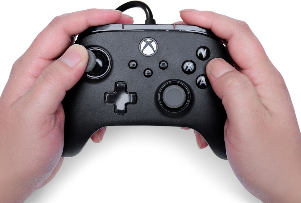 Gamepad PowerA Advantage Plus - Xbox čierny Na obrázku drží človek oboma rukami herný ovládač PowerA Advantage Plus počas hrania. Palce spočívajú na analógových páčkach a tlačidlách, čo ukazuje bežný spôsob ovládania hier. Dobre sú vidieť tlačidlá A, B, X a Y, smerový kríž, funkčné tlačidlá uprostred aj tlačidlo s logom Xbox. Snímka demonštruje ergonomický tvar ovládača, vďaka ktorému pohodlne padne do rúk aj pri dlhšom hraní.