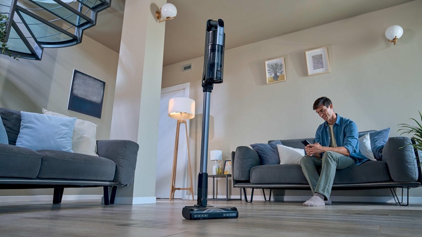 Tyčový vysávač Hoover HF310H 011 čierny/svetlo šedá Tyčový vysávač Hoover HF310H 011 stojí uprostred miestnosti, zatiaľ čo muž na gauči používa telefón