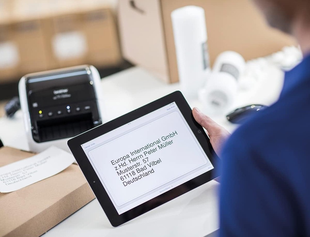 Tlačiareň štítkov Brother QL-1110NWBC čierna/biela Muž drží v ruke tablet s adresou, ktorú vytlačí v pozadí nachádzajúcej sa tlačiareň štítkov Brother QL-1110NWBC.