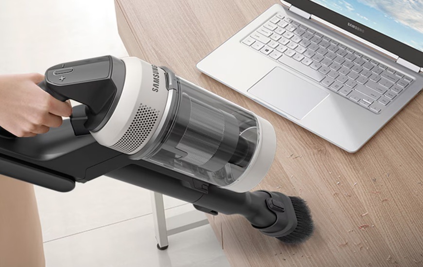 Tyčový vysávač Samsung Bespoke AI Jet VS80F28DES/GE béžový Ruka vysáva stolík, na ktorom leží notebook, vysávačom Samsung Bespoke AI Jet VS80F28DES/GE s kefou Jet Dual Brush+