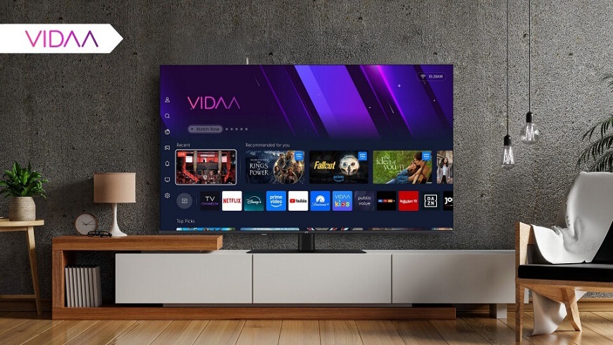 Televízia JVC na televíznom stolíku v modernom interiéri obývacej izby so zobrazením operačného systému VIDAA a stiahnutými aplikáciami