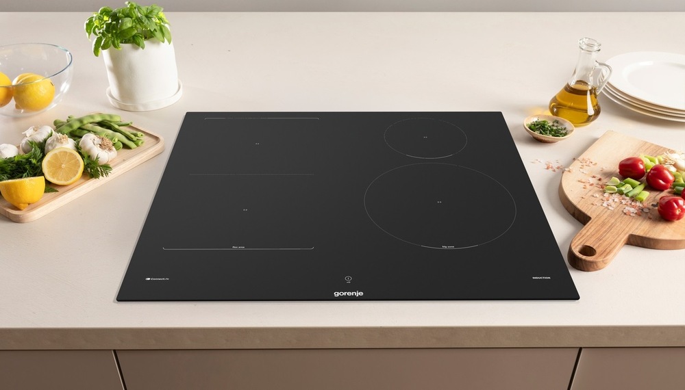 Indukčná varná doska Gorenje G800 GI6433SRWF InfiniteMatt čierna varná doska Gorenje GI6433SRWF na svetlo kuchynskej doske. Vedľa nej je ovocie a zelenina na drevených doskách