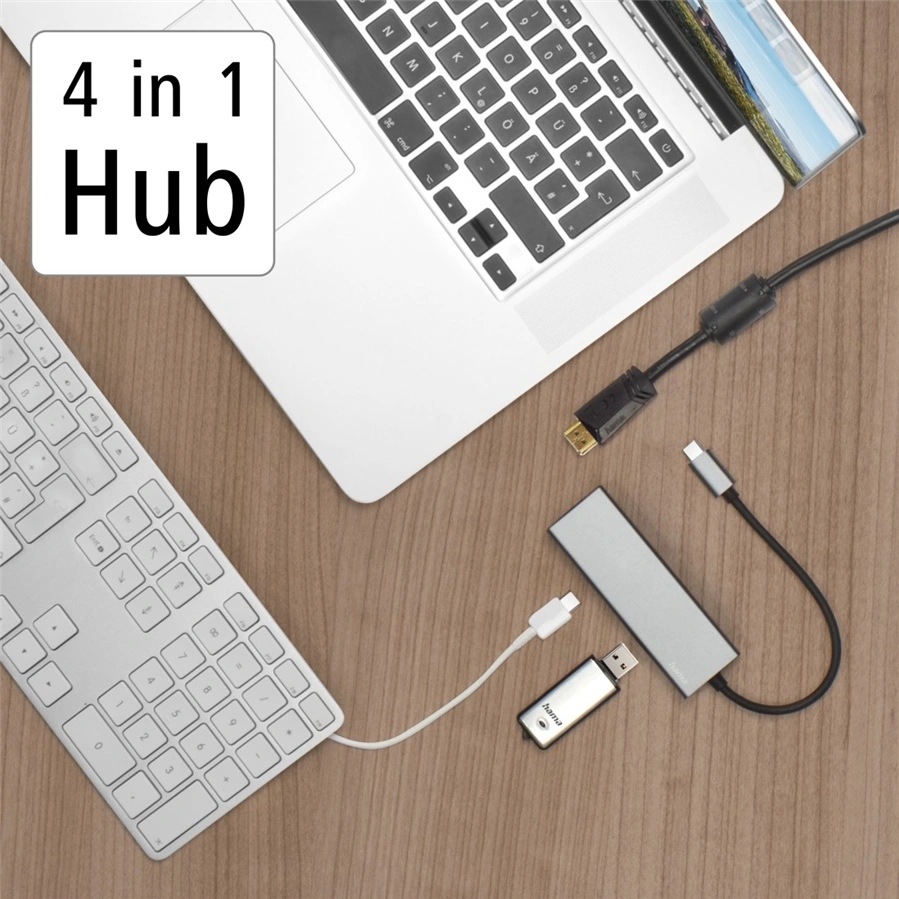 USB Hub Hama Multiport sivý Na obrázku je pracovný stôl z pohľadu zhora s notebookom, klávesnicou a šedým USB-C hubom Hama. Hub leží vpravo dole a je k nemu pripojený krátky kábel s USB-C konektorom, vedľa neho sú ďalšie káble a malý USB adaptér. Vľavo hore je výrazný nápis „4 in 1 Hub“. Fotka ukazuje praktické použitie hubu na pripojenie príslušenstva k notebooku.