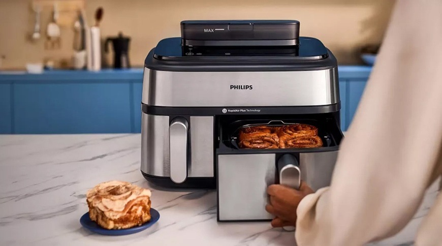 Fritéza teplovzdušná Philips Airfryer Dual Basket 5000 series 9 l, NA555/00 strieborná Žena drží madlo pri väčšom koši a vysúva ho smerom k sebe. V pozadí je vidieť zvyšok kuchynskej linky.