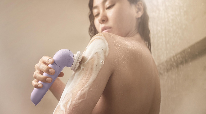 Žena používa epilátor Philips BRE728/00 vo fialovej farbe s exfoliačnou kefou na mokrej pokožke ramena, ktoré je pokryté penou počas starostlivosti o telo.