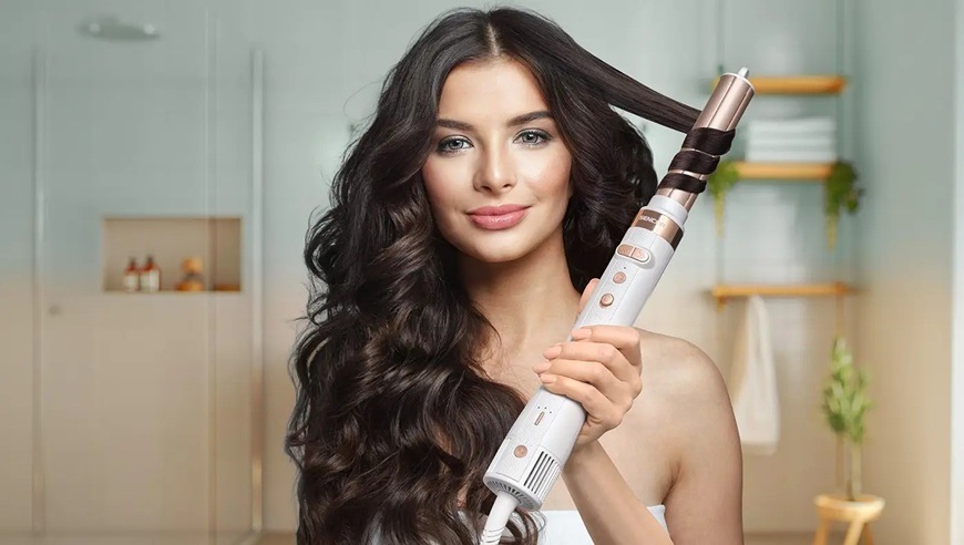 Žena s dlhými tmavými vlasmi používa multistyler Sencor SHS 9200RS s kulmovacím nástavcom, na ktorý je navinutý prameň vlasov. Prístroj má svetlé ergonomické telo s ovládacími tlačidlami a kovovým zakončením.