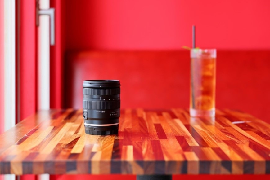 Objektív Tamron 11-20 mm F/2.8 Di III-A RXD (Canon RF-S) čierny Zátišie s nápojom a objektívom Tamron 11–20 mm F/2.8 Di III-A RXD na stolíku v kaviarni