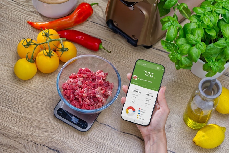 Žena drží v ruke telefón av ňom má otvorenú aplikáciu Sencor FOOD, ktorá ukazuje výživové hodnoty pripravovaného pokrmu.
