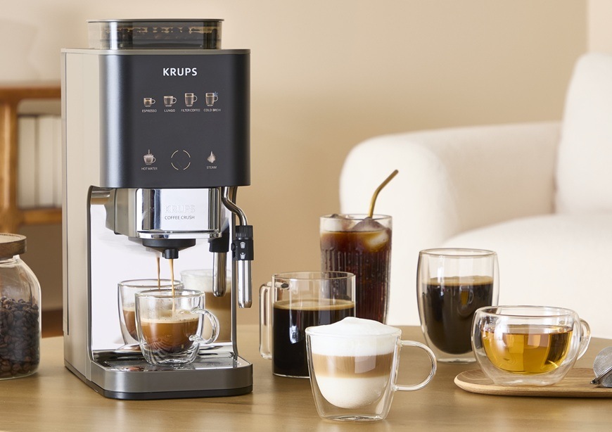 Automatický kávovar Krups Coffee Crush Experience SA403BE0 sivý/hnedý Predný pohľad na kávovar Krups SA403BE0, ktorý pripravuje horúcu vodu na čaj aj najrôznejšie druhy káv. V pozadí je biele kreslo.