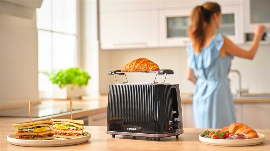Hriankovač Sencor STS 7511BK čierny V popredí je čierny hriankovač STS 7511BK položený na kuchynskej linke. Vedľa neho sú pripravené toasty a croissant. V pozadí je moderná kuchyňa. Pani zarába zo skrinky ďalší tanier.