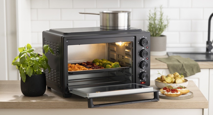 Mini rúra Gorenje OM38GBC čierna Mini rúra Gorenje OM38GBC je položená na kuchynskej linke. Jej dvierka sú pootvorené. Vnútri sa nachádza plech na pečenie so zeleninou. Na sklokeramickej doske s 2 varnými zónami je umiestnený hrniec.