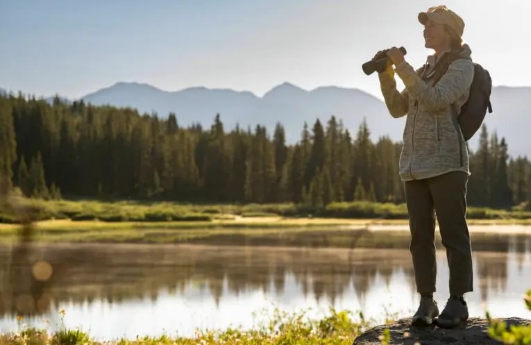 Ďalekohľad Nikon Action 10×50 čierny žena stojí s ďalekohľadom Nikon Action 10×50 v ruke na brehu rieky, okolo nej sa rozprestiera úchvatná príroda.