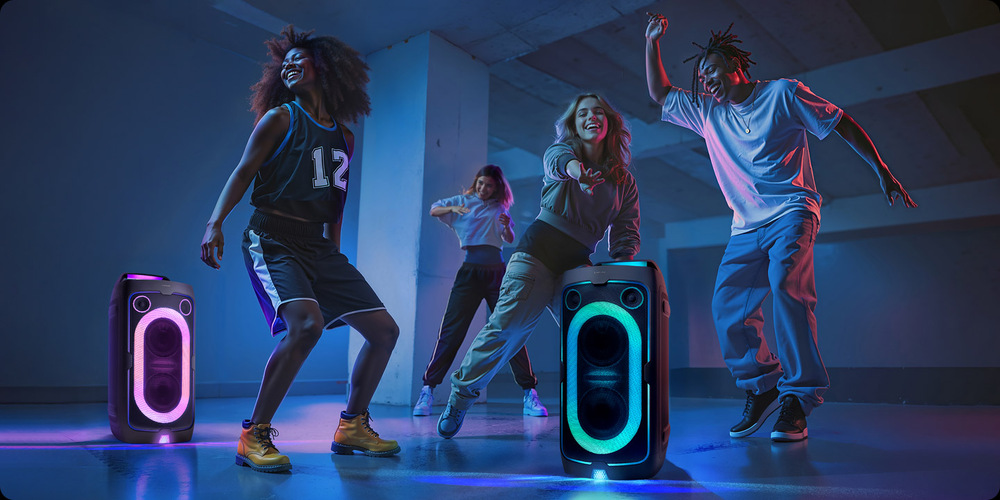 Dva reproduktory Samsung MX-ST40F stoja na zemi, jeden svieti ružovo, druhý zeleno, okolo nich tancujú štyria ľudia
