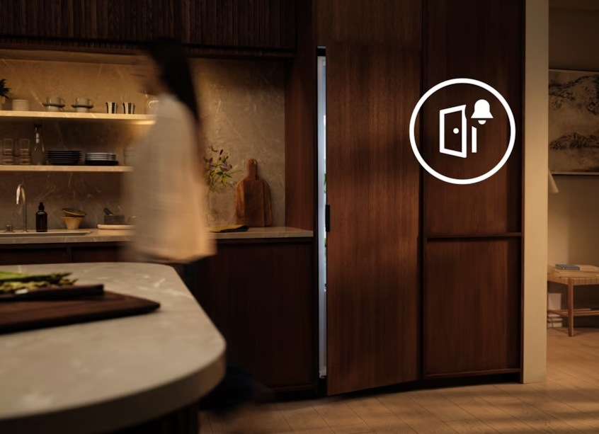 Pootvorená vstavaná chladnička Electrolux. Na skrini vedľa je symbol alarmu. Pred chladničkou je zachytená prechádzajúca žena.