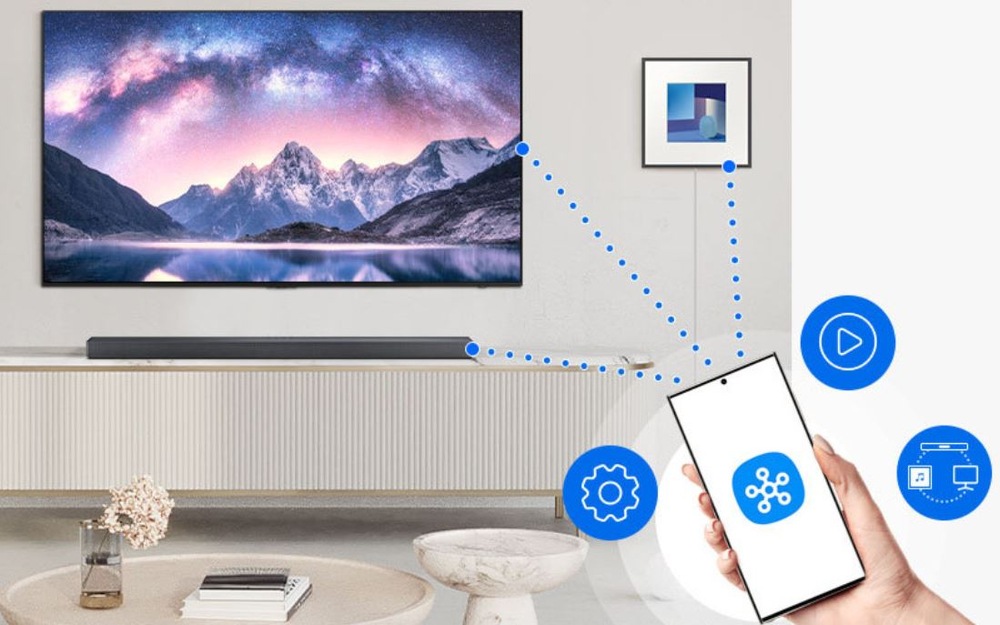 Soundbar Samsung je možné ovládať aj cez mobilnú aplikáciu. Z telefónu vychádzajú bodky k televízii a soundbaru. Televízia visí na stene a pod ňou je soundbar na stolíku.