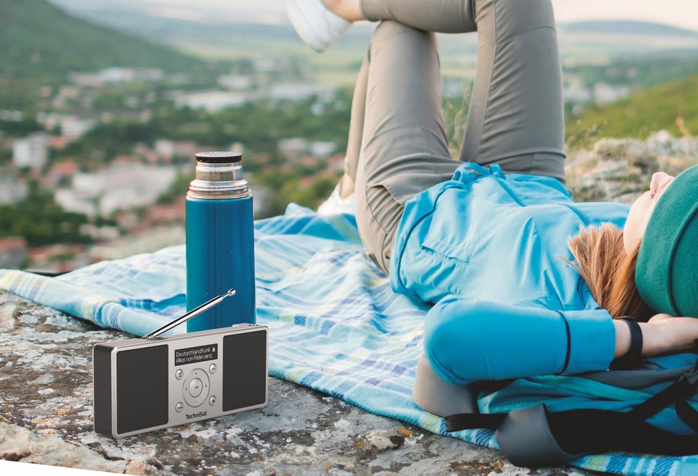 Rádioprijímač s DAB+ Technisat DIGITRADIO 1 S čierny/strieborný Rádioprijímač TechniSat DIGITRADIO 1 S s vysunutou anténou stojí na skale vedľa termosky. V pozadí odpočíva turistka na deke s výhľadom do krajiny, čo znázorňuje použitie rádia na cestách.