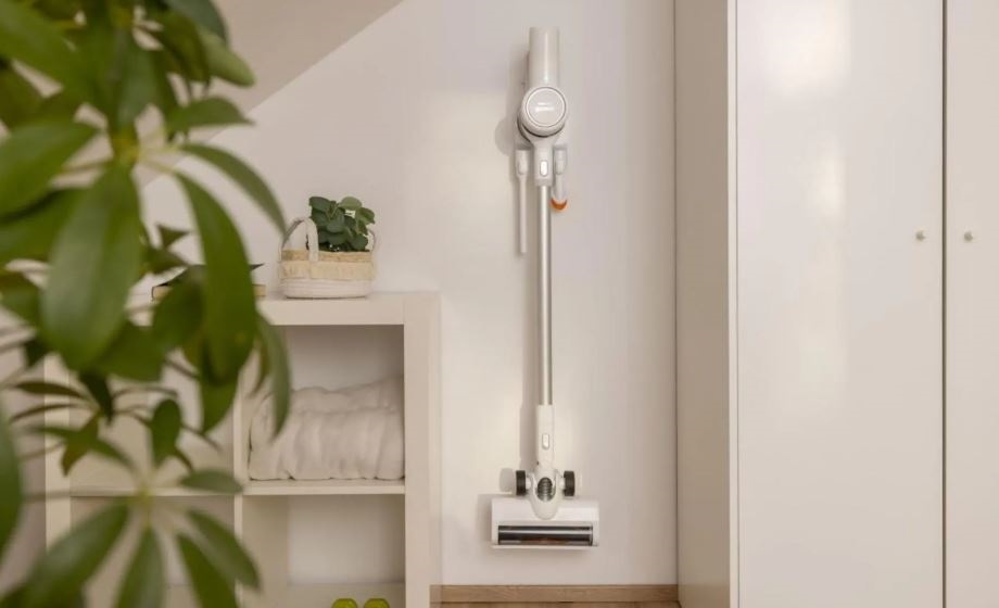 tyčový vysávač Gorenje SVC216290FFW vidí na stene vo svojom držiaku. Okolo sú rastliny a všetko je osvetlené slnkom.