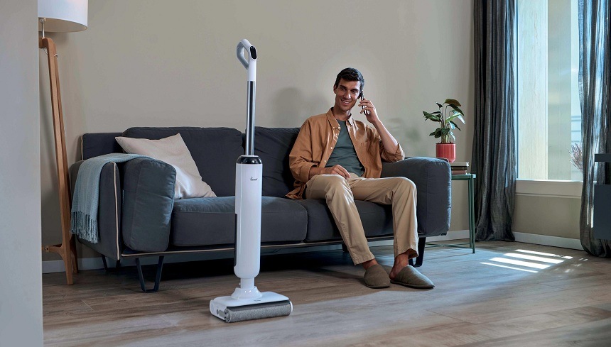 Tyčový vysávač Hoover HW400 011 biely vysávač Hoover HW400 011 stojí uprostred miestnosti, zatiaľ čo muž sedí a telefonuje na gauči