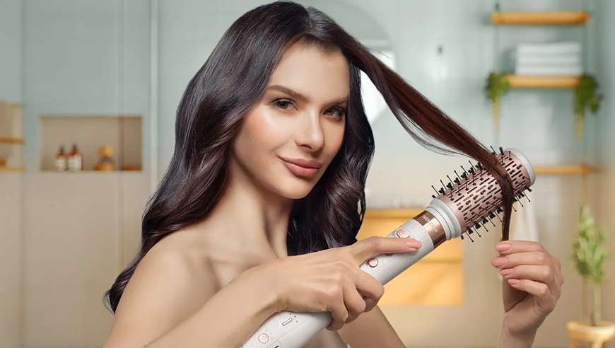 „Žena s dlhými tmavými vlasmi používa multistyler Sencor SHS 9200RS s okrúhlym kefovým nástavcom, okolo ktorého je omotaný prameň vlasov. Jednou rukou drží prístroj s ovládacími tlačidlami, druhou upravuje vlasy.