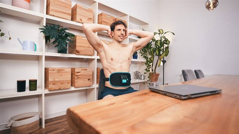 Elektrostimulátor Beurer Corefit II čierny Muž sedí v izbe pri stole a používa brušný pás Beurer Corefit II