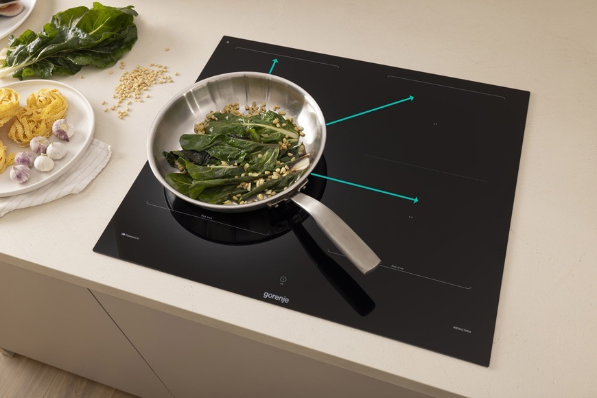 Indukčná varná doska Gorenje GI6443BSCWF je zabudovaná v modernej kuchyni. Na doske sa v panvici pripravuje jedlo. Modré šípky graficky ukazujú, že panvicu je možné presúvať na iné varné zóny bez straty nastavenia výkonu a času.