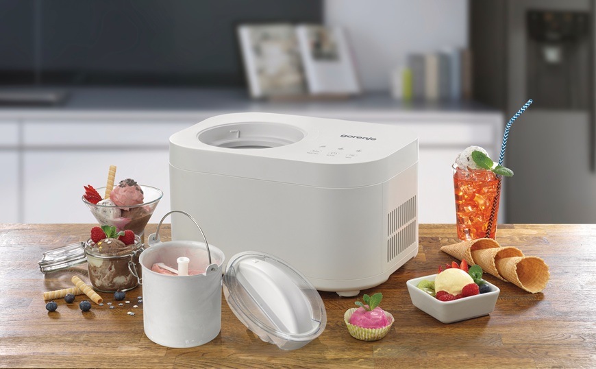 Zmrzlinovač Gorenje ICM10W je položený na kuchynskej doske. Okolo neho je zmrzlinový pohár s ovocím a chladený drink.