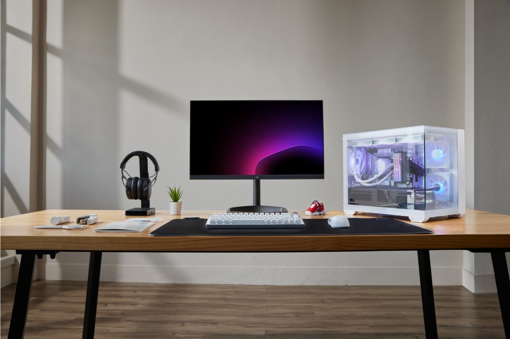 Moderná počítačová zostava na drevenom stole obsahuje elegantný monitor s fialovým gradientným pozadím, biely herný počítač, klávesnicu, myš, slúchadlá a malú rastlinu.