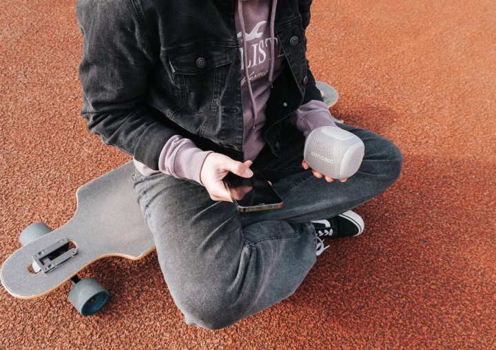 Užívateľ drží reproduktor Concept SoundJoy BR5011 a telefón pri posedení vedľa longboardu