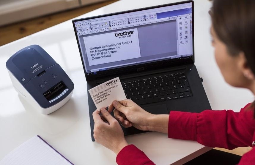 Tlačiareň štítkov Brother QL-600 biela/modrá žena sedí pri počítači, na ktorom pripravila štítok podľa požiadaviek, vedľa je na stole položená aj tlačiareň štítkov Brother QL-600 s vytlačenou etiketou.