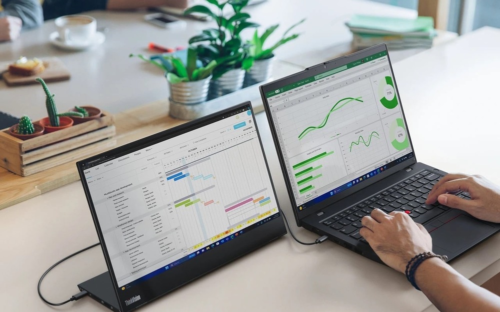 Notebook Lenovo ThinkPad L14 Gen 6 čierny Na stole sú dva notebooky, na ktorých sú zobrazené grafy a plány, obklopené malými rastlinami v kvetináčoch, a osoba, ktorá píše na klávesnici, vytvára sústredenú pracovnú atmosféru.