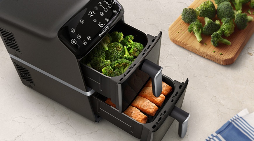 Fritéza teplovzdušná Philips Airfryer Dual Basket 4000 series 10 l, NA461/00 čierna Teplovzdušná fritéza Philips NA461/00 s dvoma vysunutými košmi, v hornom koši je brokolica av spodnom filety lososa, vedľa na kuchynskej linke je drevená doštička s ďalšou zeleninou.