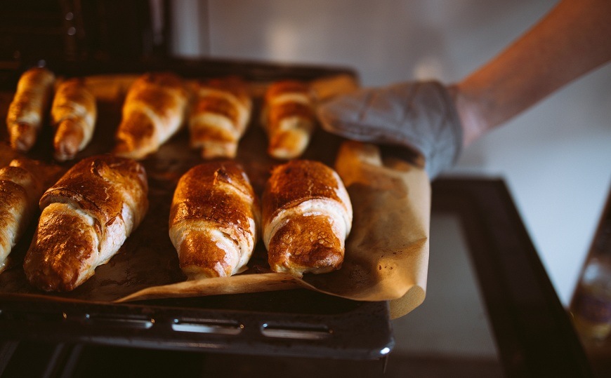 Ruka v šedej kuchynskej chňapke vyťahuje z rúry ETA 4786 plech plný čerstvo upečených zlatistých croissantov. Pečivo leží na papieri na pečenie a je rovnomerne prepečené, s lesklým povrchom